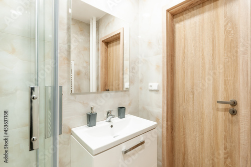 Contemporary bathroom with glassy walled shower and sink with white cabinet under it and mirror on wall. All walls are lined with marble in beige shades. Door are made of wood.