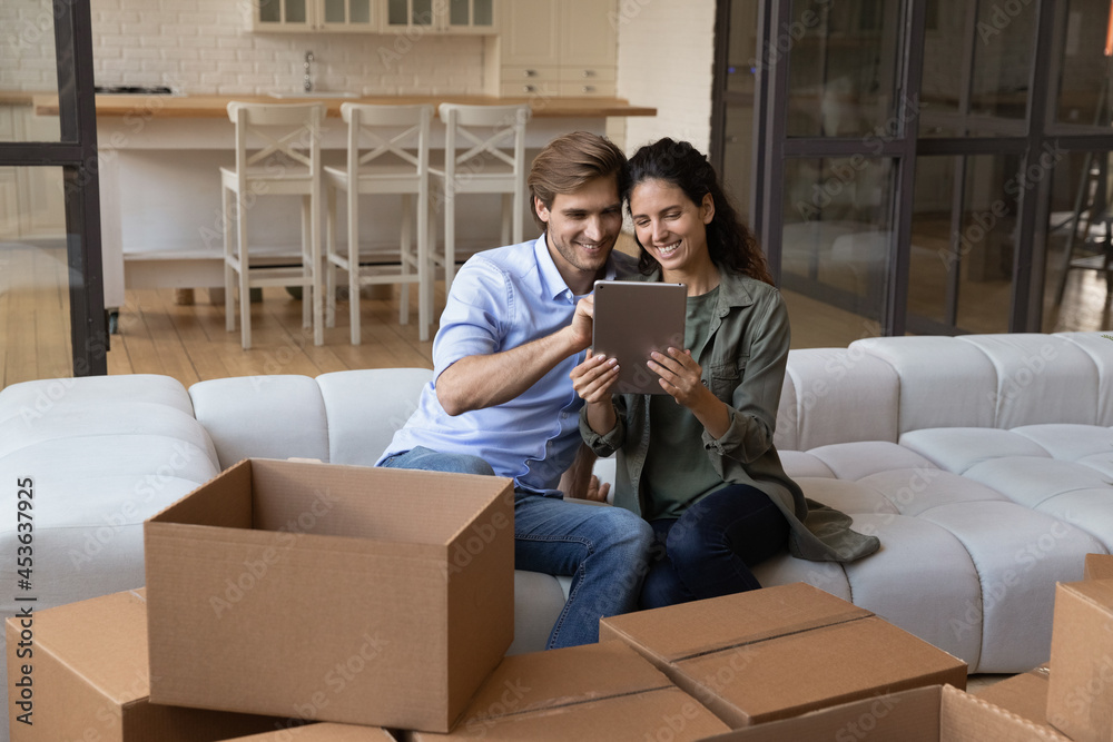 Happy young family couple using digital computer tablet, sitting on sofa near huge boxes, purchasing decorations for renovated living room in internet store or leaving review on real estate agency. Happy young family couple using digital computer tablet, sitting on sofa near huge boxes, purchasing decorations for renovated living room in internet store or leaving review on real estate agency.