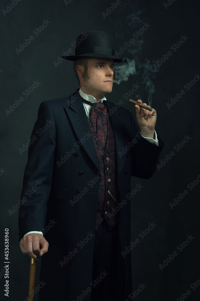 Cigar smoking young man in stylish vintage Victorian clothes with a walking stick stands in a room in front of a dark gray wall.