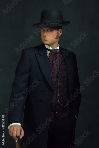 Young man in stylish vintage victorian attire. Standing with a cane in front of a dark gray wall.