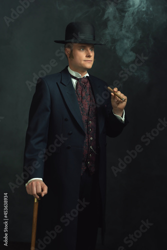 Cigar smoking young man in stylish vintage Victorian clothes with a walking stick stands in a room in front of a dark gray wall.