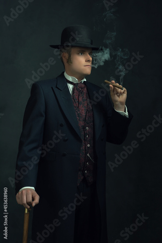 Obraz na płótnie Cigar smoking young man in stylish vintage Victorian clothes with a walking stick stands in a room in front of a dark gray wall. w salonie