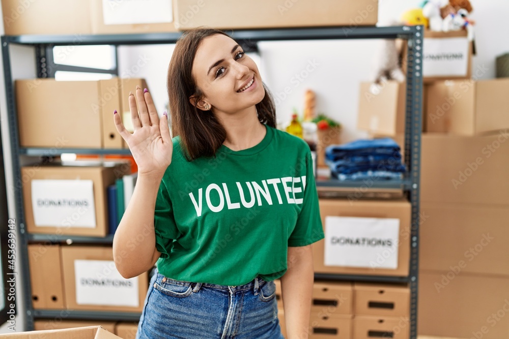 Young brunette woman wearing volunteer t shirt at donations stand waiving saying hello happy and smiling, friendly welcome gesture Young brunette woman wearing volunteer t shirt at donations stand waiving saying hello happy and smiling, friendly welcome gesture