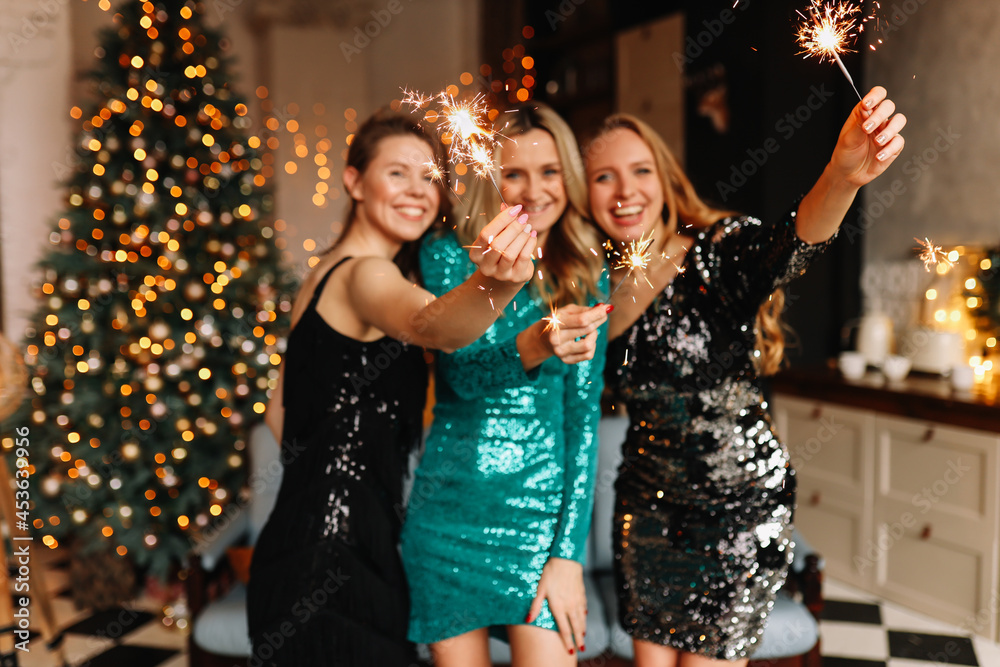 Funny girls friends in Santa hats celebrate the Christmas holiday laugh rejoice hold glasses drink champagne burn sparklers at a party in the decorated kitchen in the loft style of the house