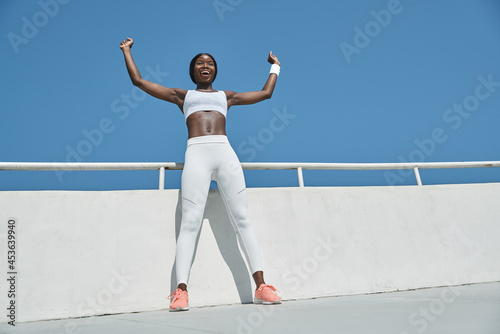 Low angle view of beautiful young African woman in sports clothing keeping arms raised and smiling