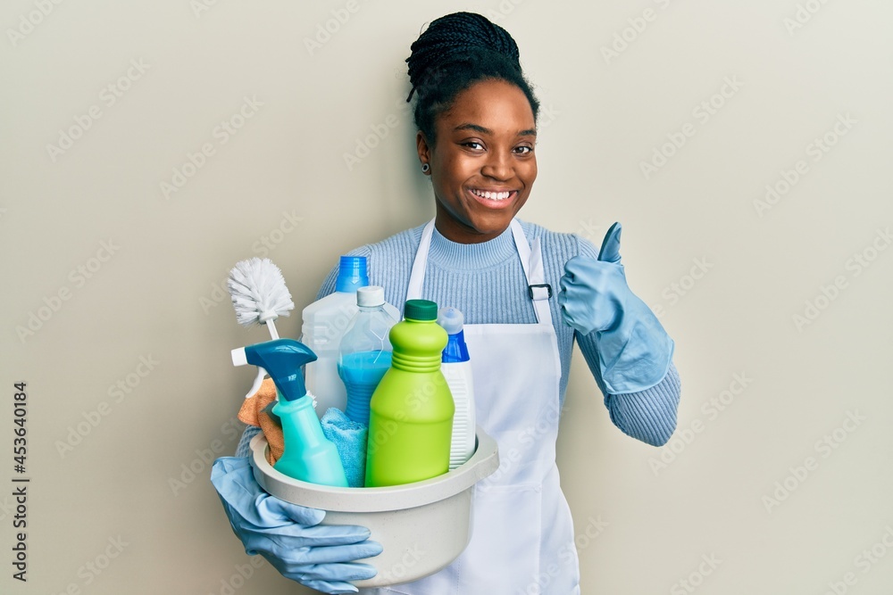 African american woman with braided hair wearing apron holding cleaning products smiling happy and positive, thumb up doing excellent and approval sign African american woman with braided hair wearing apron holding cleaning products smiling happy and positive, thumb up doing excellent and approval sign