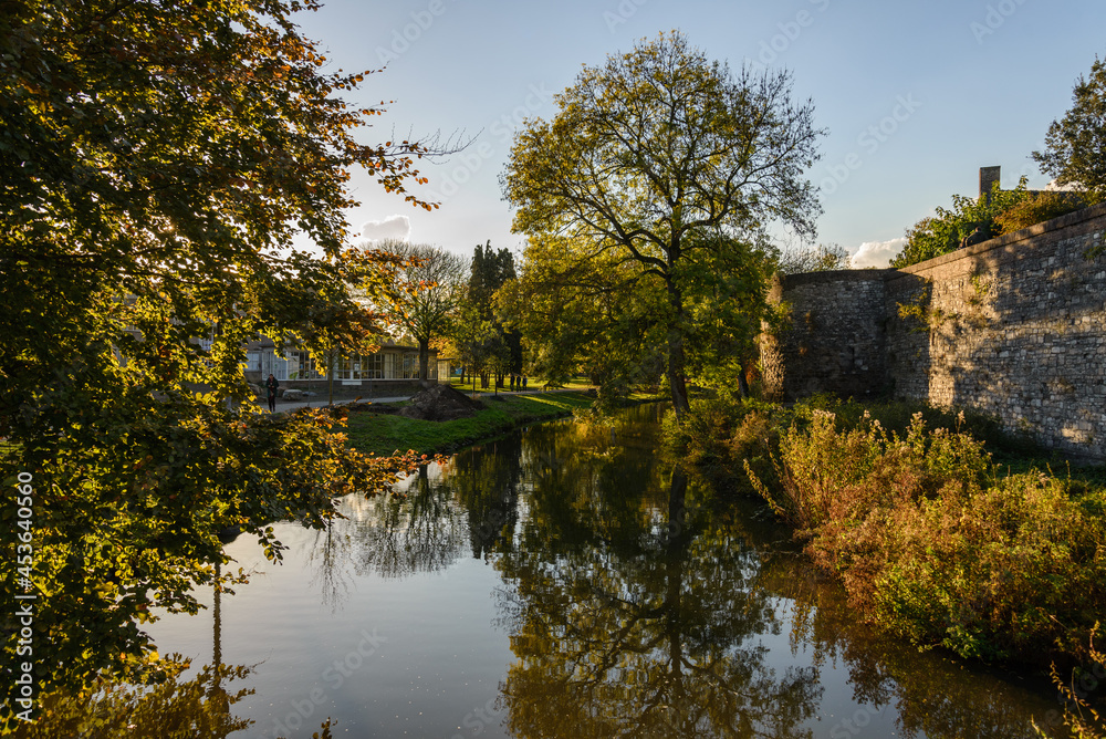 Outdoor scenery of walking way along Jeker canal and historical city wall at Monseigneur Nolenspark, city public park, in Autumn season, in Maastricht, Netherlands during evening sunset time.
