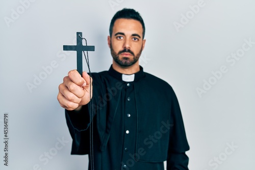 Handsome hispanic priest man with beard holding catholic cross thinking attitude and sober expression looking self confident