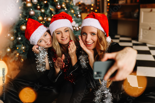 Happy funny girls girlfriends in Santa hats celebrate Christmas holiday laugh take photo selfies using mobile phone technology at a party at home