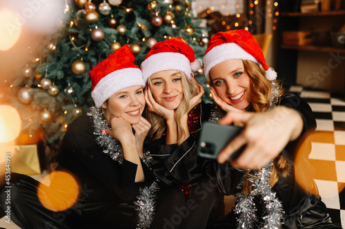 Happy funny girls girlfriends in Santa hats celebrate Christmas holiday laugh take photo selfies using mobile phone technology at a party at home