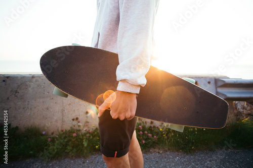 Young hipster man holds cool longboard in hand. Sun sunset light leak in lens. Beautiful travel wanderlust concept. Millennial adventure trip. Skater walk with skateboard 