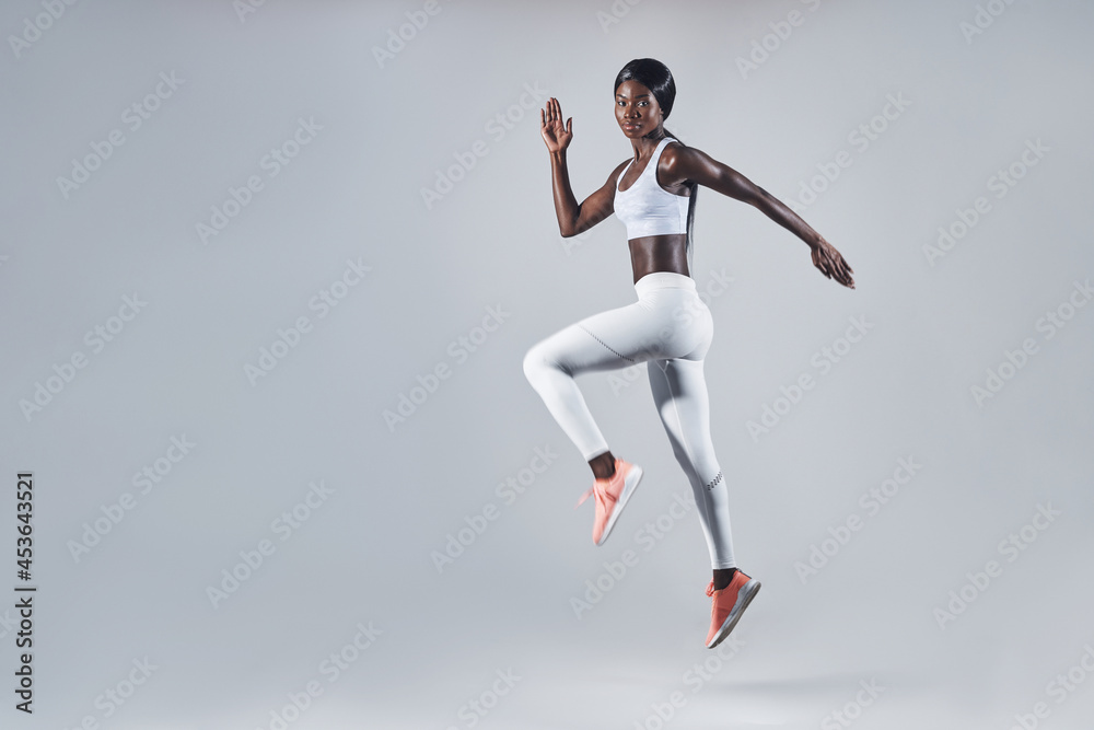 Full length of confident young African woman in sports clothing jumping against gray background