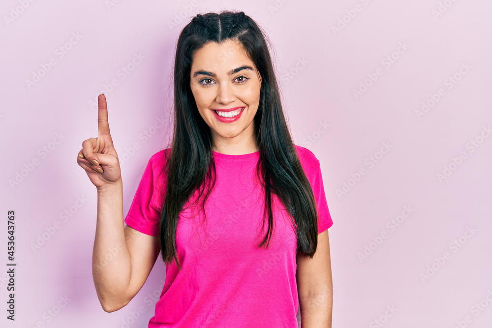 Young hispanic girl wearing casual pink t shirt showing and pointing up with finger number one while smiling confident and happy. Young hispanic girl wearing casual pink t shirt showing and pointing up with finger number one while smiling confident and happy.