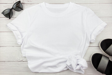 White womens cotton T Shirt mockup with summer shoes sandals and sunglasses on white wooden background. Design t shirt template, print presentation mock up. Top view flat lay.