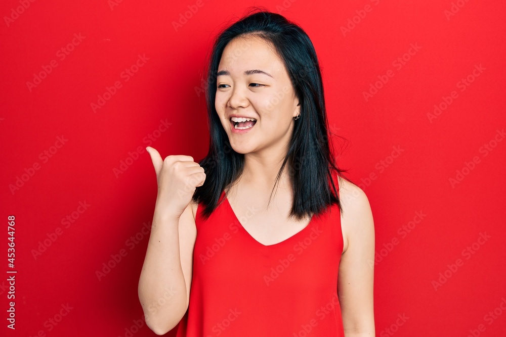 Young chinese girl wearing casual clothes pointing thumb up to the side smiling happy with open mouth Young chinese girl wearing casual clothes pointing thumb up to the side smiling happy with open mouth