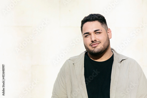 Fat latin man, looking at the camera, with a cute and friendly expression. With a white wall of the street in the background. Latins and curvy models concept.