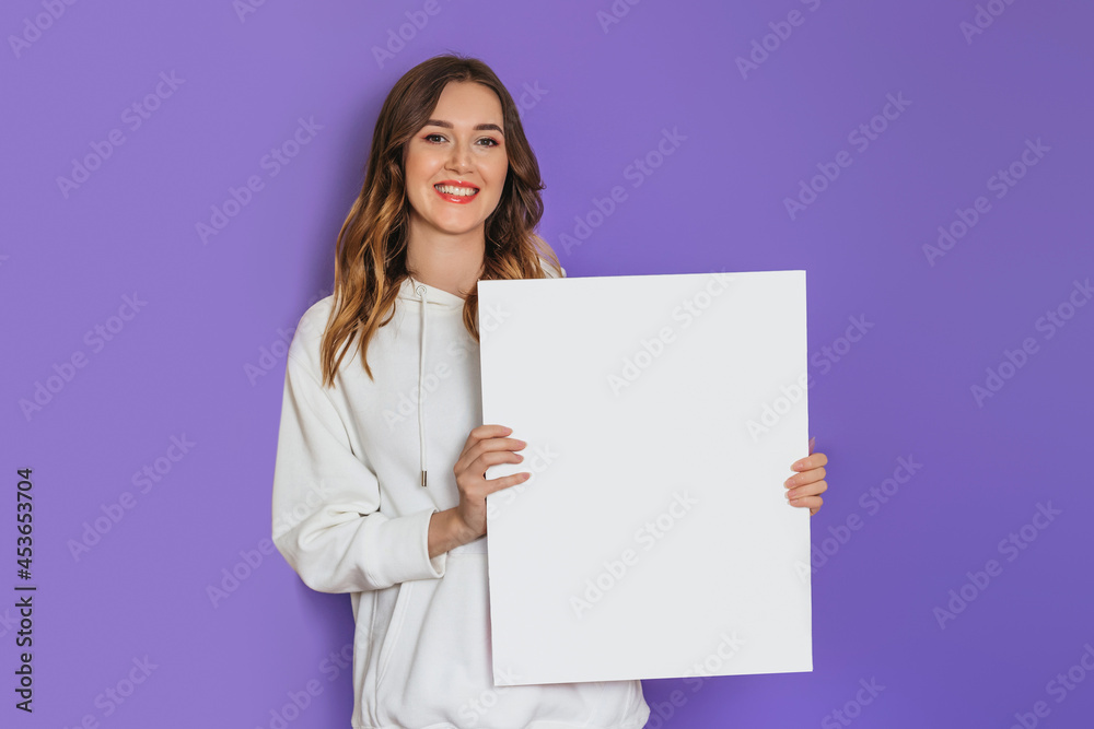 Young caucasian student girl holding a white sheet of paper, poster, placard in hands smiling isolated on lilac background Young caucasian student girl holding a white sheet of paper, poster, placard in hands smiling isolated on lilac background