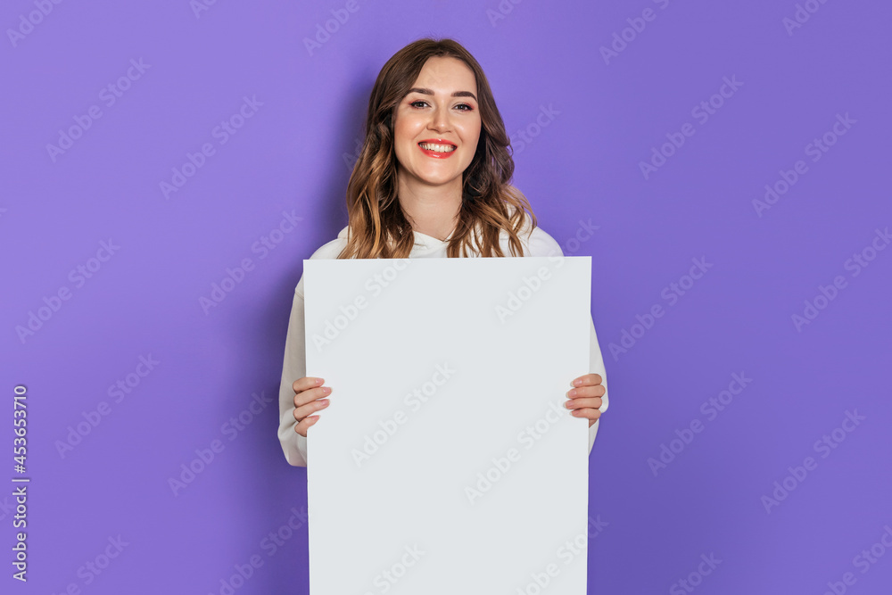 Young caucasian student girl holding a white sheet of paper, poster, placard in hands smiling isolated on lilac background. Copy space Young caucasian student girl holding a white sheet of paper, poster, placard in hands smiling isolated on lilac background. Copy space