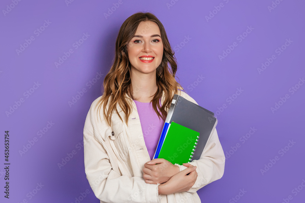 Caucasian student girl holding a book, notebook smiling, looking at the camera isolated on lilac background in studio Caucasian student girl holding a book, notebook smiling, looking at the camera isolated on lilac background in studio