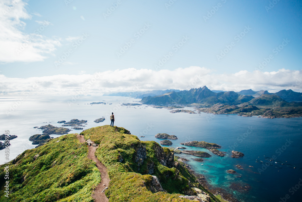Amazing epic shot of young man hiker stand on top of mountain after long difficult hike in Lofoten Islands in Norway. Incredible summer views of scandinavian travel lifestyle. Epic landscape in north - obrazy, fototapety, plakaty 