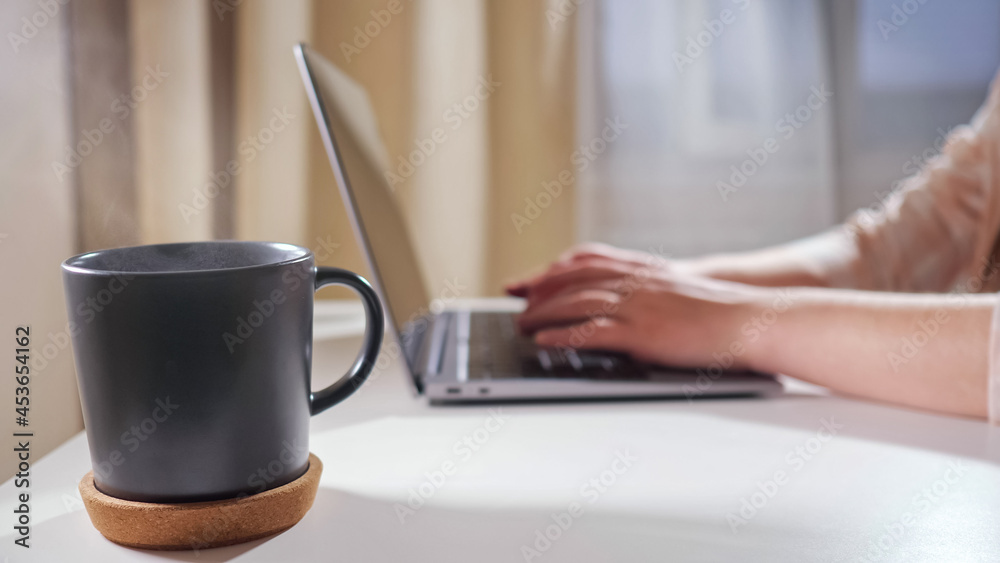Close-up of a mug with a hot drink on the table against the background of an unrecognizable woman typing on a laptop. Close-up of a mug with a hot drink on the table against the background of an unrecognizable woman typing on a laptop.
