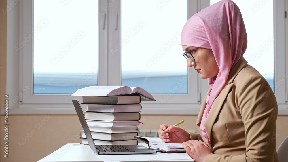 Muslim woman writes with a pen in a notebook looking into a laptop. Muslim woman writes with a pen in a notebook looking into a laptop.