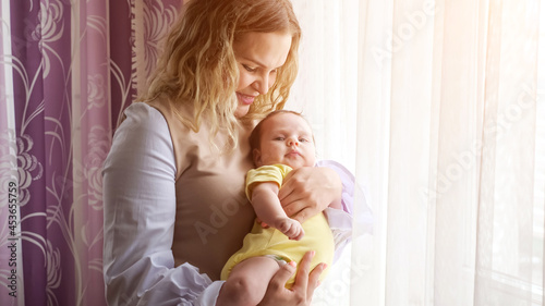 Happy young mother with curly loose hair holds in arms baby girl in yellow bodysuit kissing near window with colorful curtains in apartment room  sunlight
