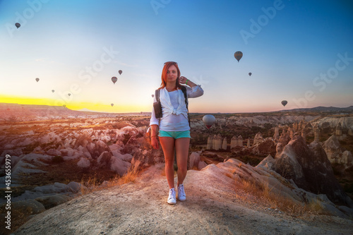 Girl with a backpack posing against the backdrop of a valley with mountains and balloons at sunrise. Entertainment, tourism an vacation. Travel tour. Goreme, Cappadocia, Turkey. © Sergey