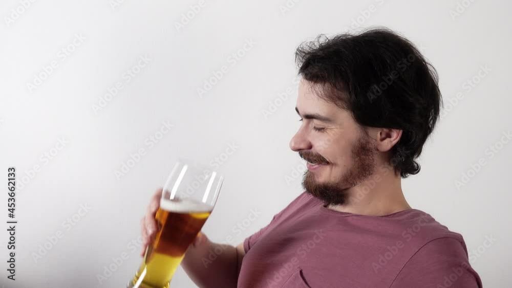 Smiling man is sitting on white background and drinking golden beer from glass talking with somebody. Profile view of a male bearded person enjoying alcohol beverage and nice discussion with a friend. Smiling man is sitting on white background and drinking golden beer from glass talking with somebody. Profile view of a male bearded person enjoying alcohol beverage and nice discussion with a friend.