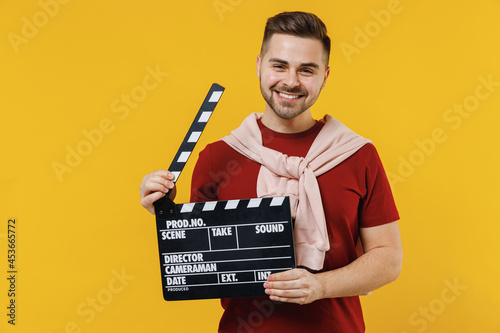 Smiling fun happy cheerful young caucasian man 20s wearing red t-shirt casual clothes holding classic black film making clapperboard isolated on plain yellow color wall background studio portrait