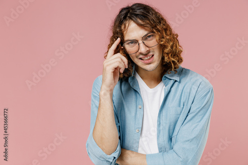 Young confused minded man 20s with long curly hair wearing blue shirt white t-shirt glasses prop up forehead with index finger head isolated on pastel plain pink color wall background studio portrait.
