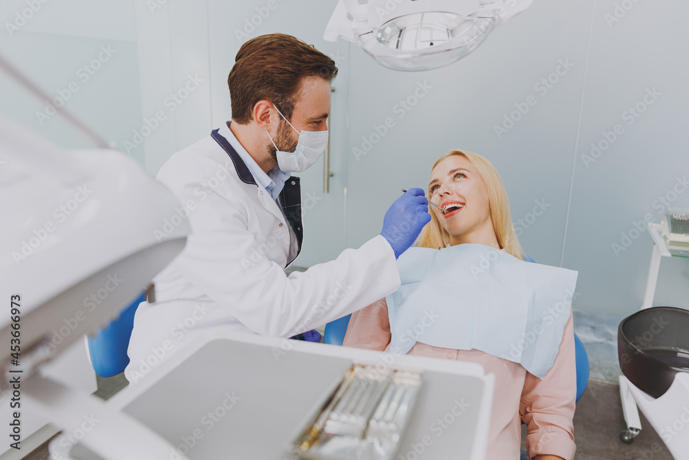 Young caucasian man doctor in white gown mask make oral examination for patient woman sitting at dentist office chair indoor cabinet near stomatologist wait medical cure Healthcare enamel treatment