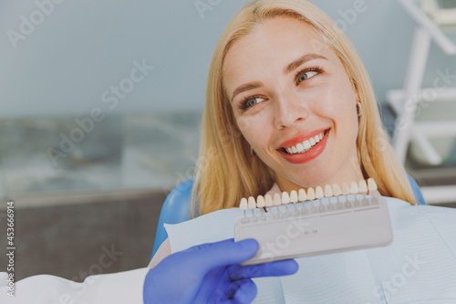 Close up young smiling european woman 20s looking at veneers enamel color palette sit at dentist office chair indoor light modern cabinet near stomatologist Healthcare oral enamel aesthetic treatment