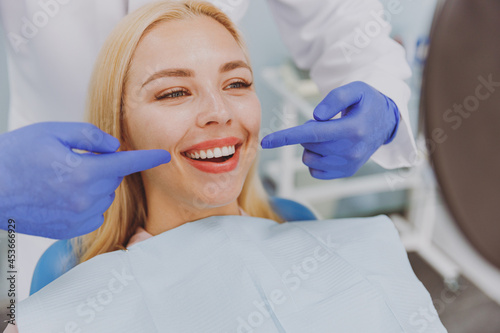 Close up young satisfied happy satisfied fun woman 20s with toothy smile looking at mirror stomatologist showing result sit at dentist office chair indoor modern cabinet Oral enamel caries treatment.