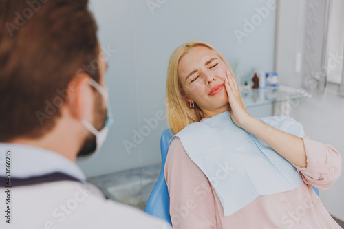 Young sick woman 20s looking at dentist sit at office chair indoor light cabinet hold cheeck visiting stomatologist with toothache painful problem wait for cure Healthcare oral enamel caries treatment