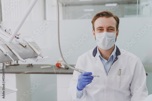 Young specialist smiling happy professional stomatologist cauasian doctor man 20s wear white gown holding show tooth drilling machine at dentist office indoor cabinet. Healthcare aesthetic treatment