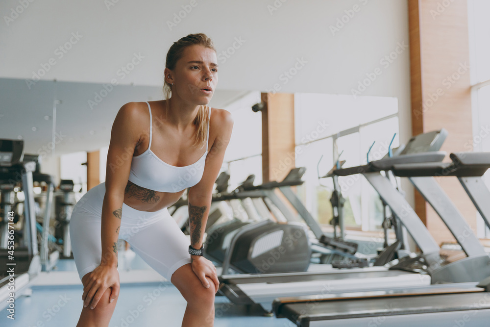 Young tired strong skinny sporty athletic sportswoman woman 20s wear white sportswear warm up training near treadmill stand leaning on knees breathing in gym indoor Workout sport motivation concept.