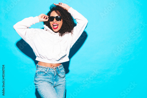 Beautiful black woman with afro curls hairstyle.Smiling model in trendy summer clothes. Sexy carefree female posing near blue wall in studio.In sunglasses with red lips