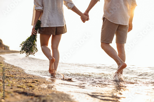 Cropped close up back view young couple two family man woman in white clothes hold flowers bouquet hand walk in water waves at sunrise over sea beach ocean outside seaside in summer day sunset evening © ViDi Studio