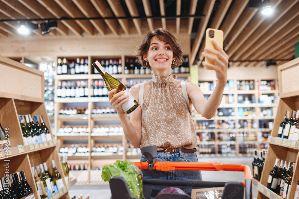 Young happy woman in casual clothes shopping at supermaket store with grocery cart hold white wine alcohol hold bottle do selfie shot on mobile phone inside hypermarket Purchasing gastronomy concept.