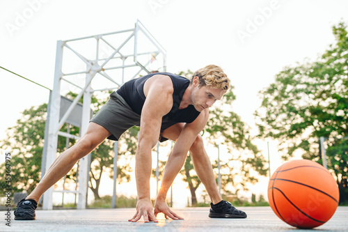Full length young sporty muscular sportsman man 20s wearing sports clothes training do stretch exercise for legs play with ball at basketball game playground court. Outdoor courtyard sport concept © ViDi Studio