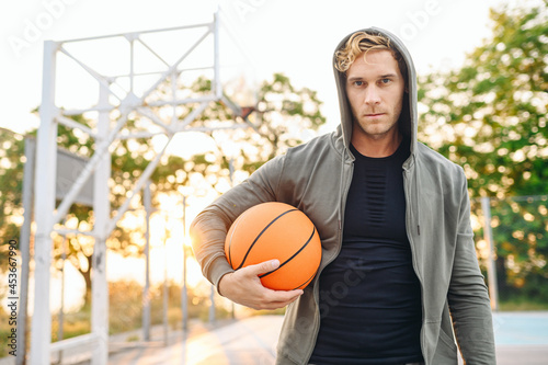 Serious cool confident sunlit young sporty sportsman man in grey sports clothes hood look camera training hold in hand ball play at basketball game playground court. Outdoor courtyard sport concept © ViDi Studio