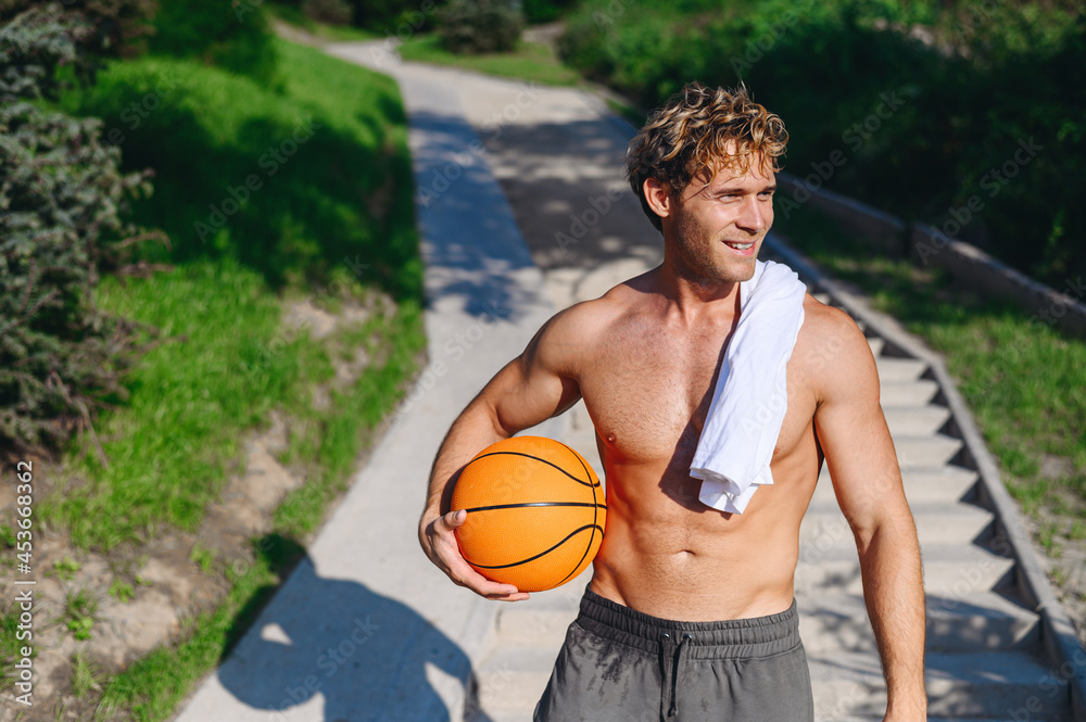Young happy strong muscular sportsman man with naked torso wear shorts training hold basketball ball waving hand walking jog run on steps up stairs in morning park on resort Helthy lifestyle concept