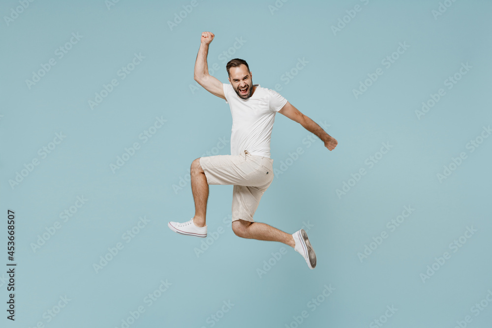 Full length side view young overjoyed happy fun excited man 20s wear casual white t-shirt jump high do winner gesture clench fist isolated on plain pastel light blue color background studio portrait