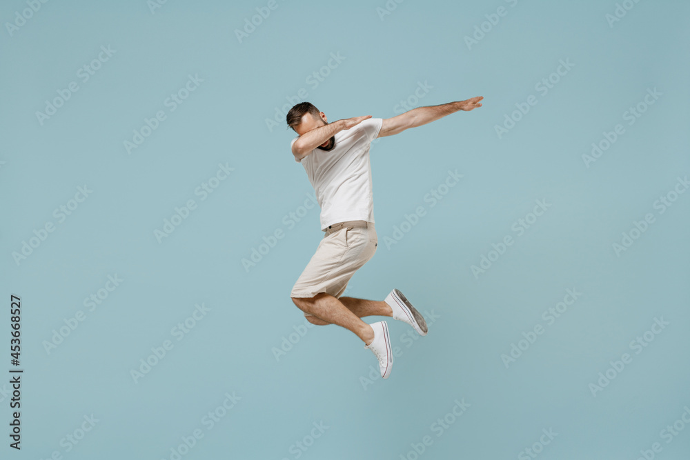 Full length young man 20s in casual white t-shirt jump high doing dab hip hop dance hands move gesture youth sign hide cover face isolated on plain pastel light blue color background studio portrait