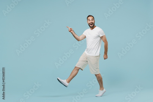 Full length side view young surprised happy man in casual white t-shirt walk going point index finger aside on workspace area copy space mock up isolated on plain pastel light blue background studio © ViDi Studio