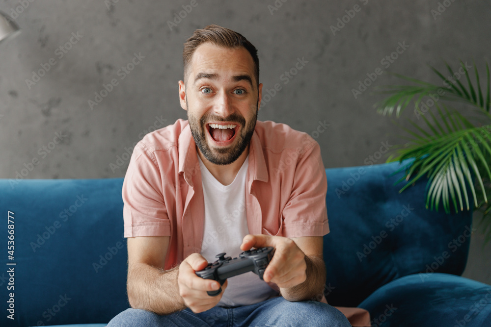 Young excited happy man in casual clothes hold in hand play pc game with joystick console sitting on blue sofa at home flat indoors rest relax on weekends free time. People lounge lifestyle concept