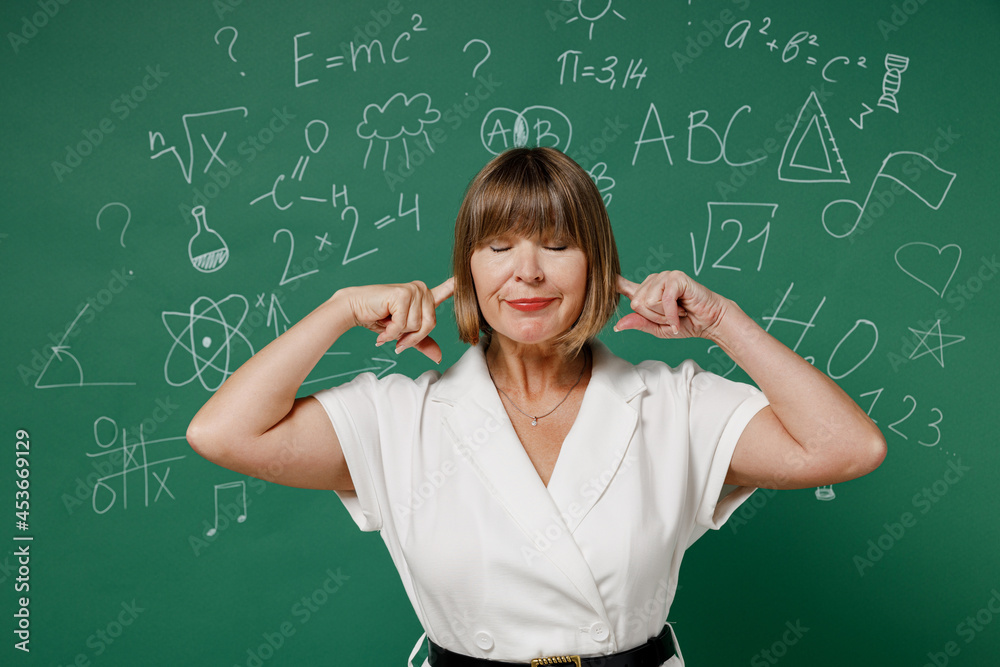 Strict teacher mature elderly senior elegant lady woman 55 wear white shirt closed eyes cover ears with hands fingers do not want to listen isolated on green wall chalk blackboard background studio