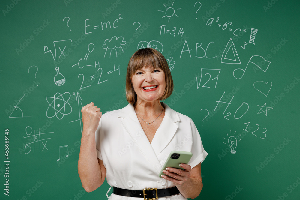 Happy excited glad teacher mature elderly senior lady woman 55 wear white shirt hold in hand use mobile cell phone clenching fists say yes isolated on green wall chalk blackboard background studio.