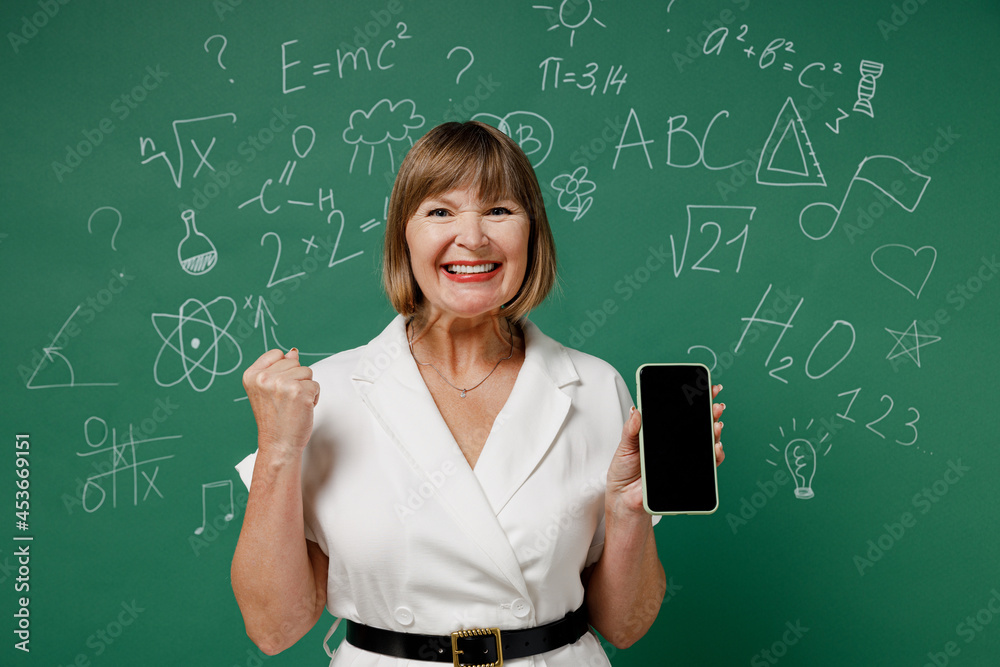 Teacher mature elderly woman 55 wear white shirt hold in hand use mobile cell phone with blank screen workspace area clenching fist say yes isolated on green wall chalk blackboard background studio